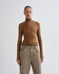 A model wearing a fitted, long-sleeve brown ruffled mock neck top and beige high-waisted jeans, standing against a light gray studio background.