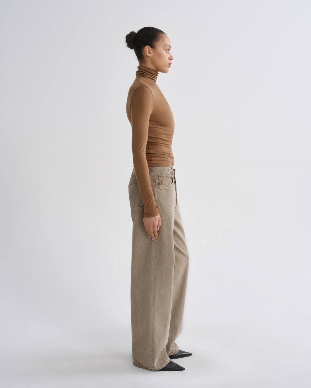 A woman in side profile wearing a fitted brown turtleneck and wide-leg beige trousers, standing against a plain light gray studio background.