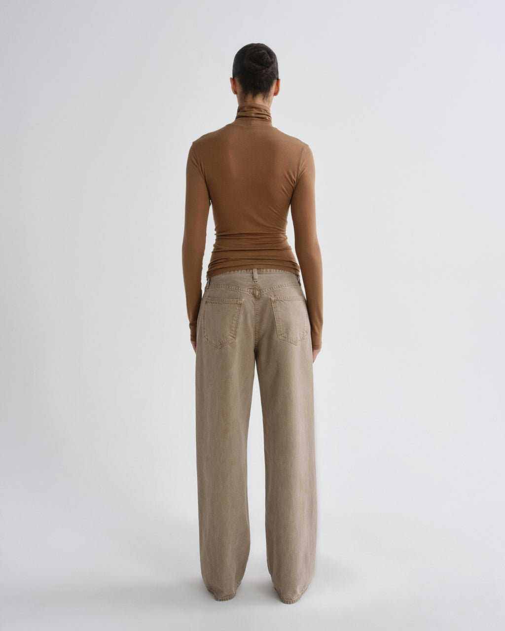 A model shown from the back wearing a brown ribbed mock neck top and beige wide-leg trousers, standing against a light gray studio background.