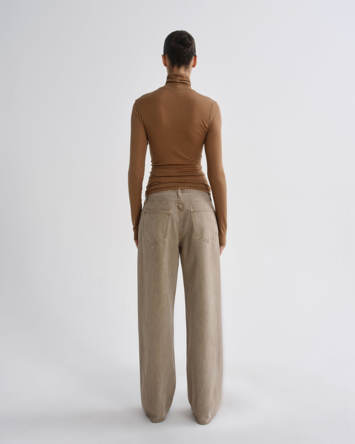 A model shown from the back wearing a brown ribbed mock neck top and beige wide-leg trousers, standing against a light gray studio background.
