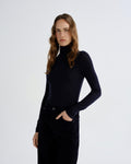 A woman modeling a fitted black long-sleeve mock neck top, paired with black corduroy trousers, against a plain light gray studio background.