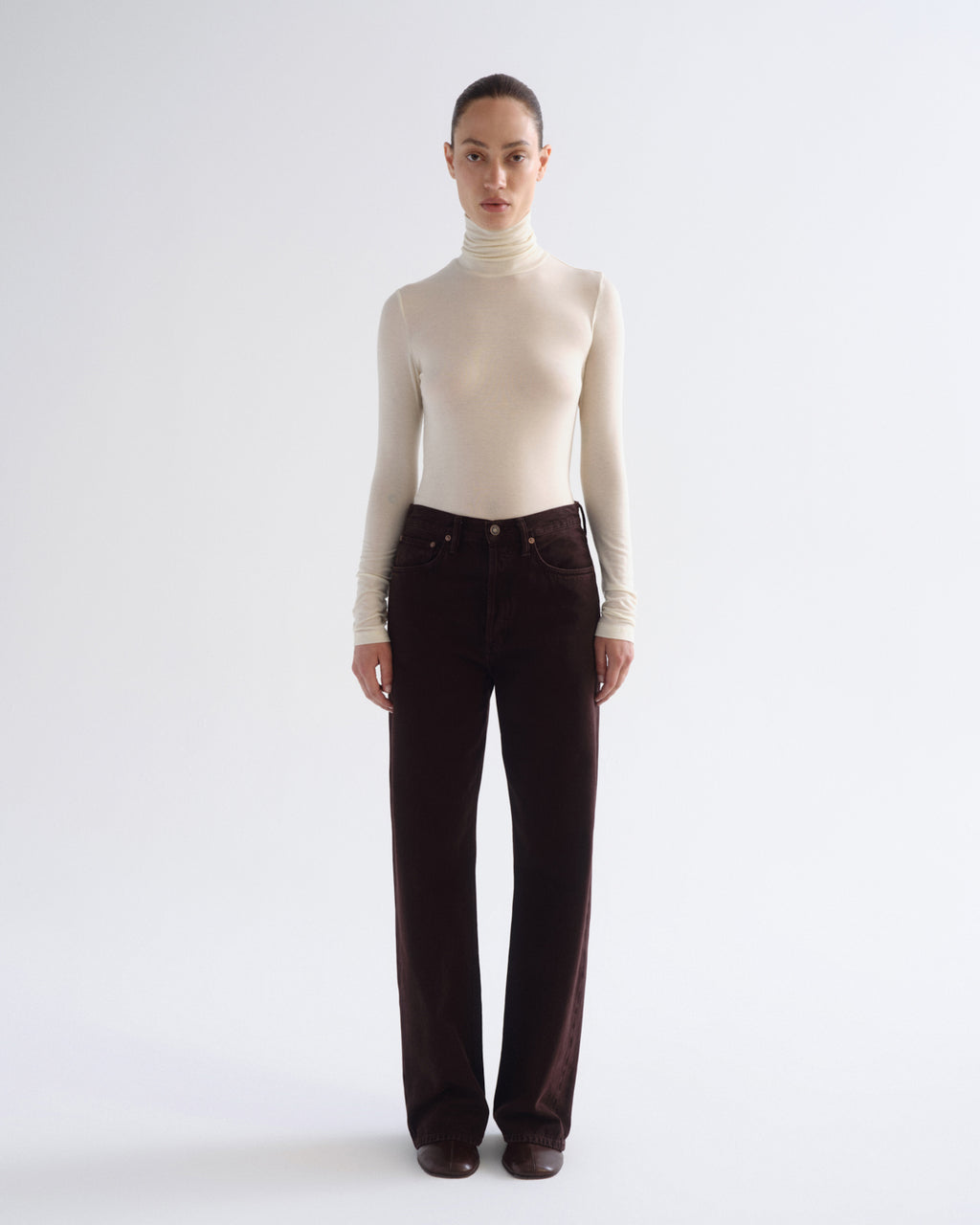 A model stands against a light gray studio background wearing a fitted cream turtleneck sweater and dark brown wide-leg corduroy pants.