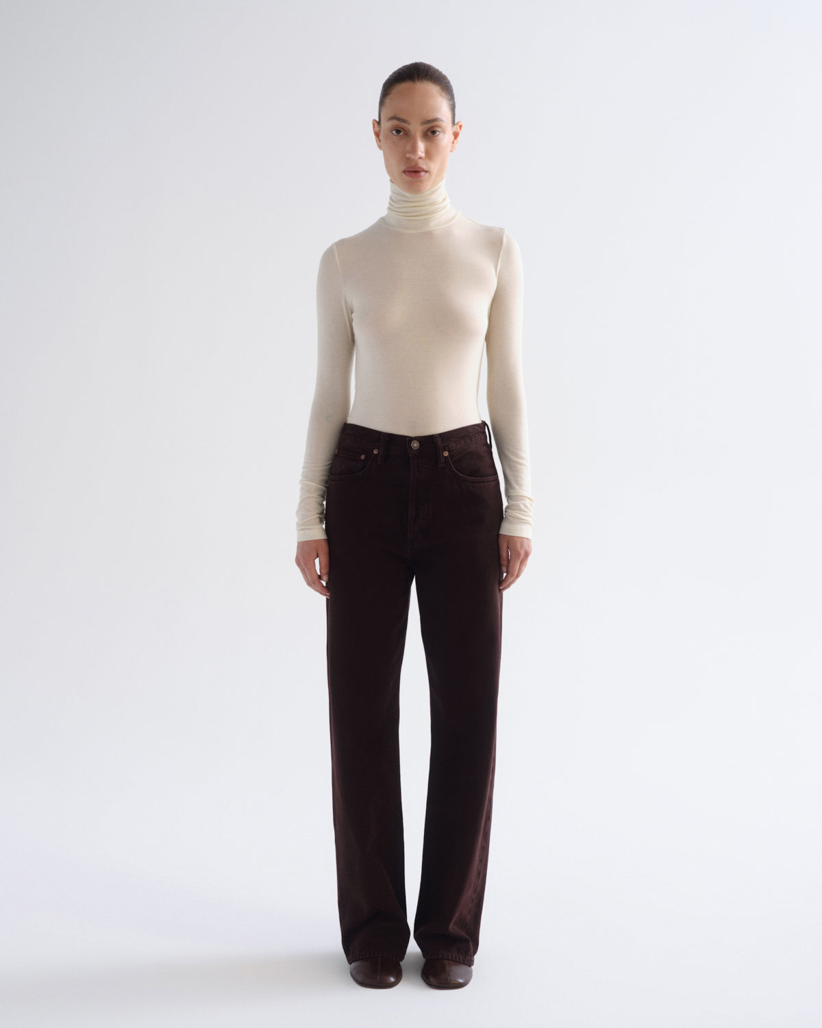 A model stands against a light gray studio background wearing a fitted cream turtleneck sweater and dark brown wide-leg corduroy pants.