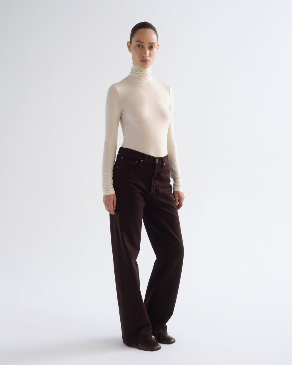 A model stands against a light gray studio background wearing a fitted cream turtleneck top and dark brown wide-leg corduroy pants.