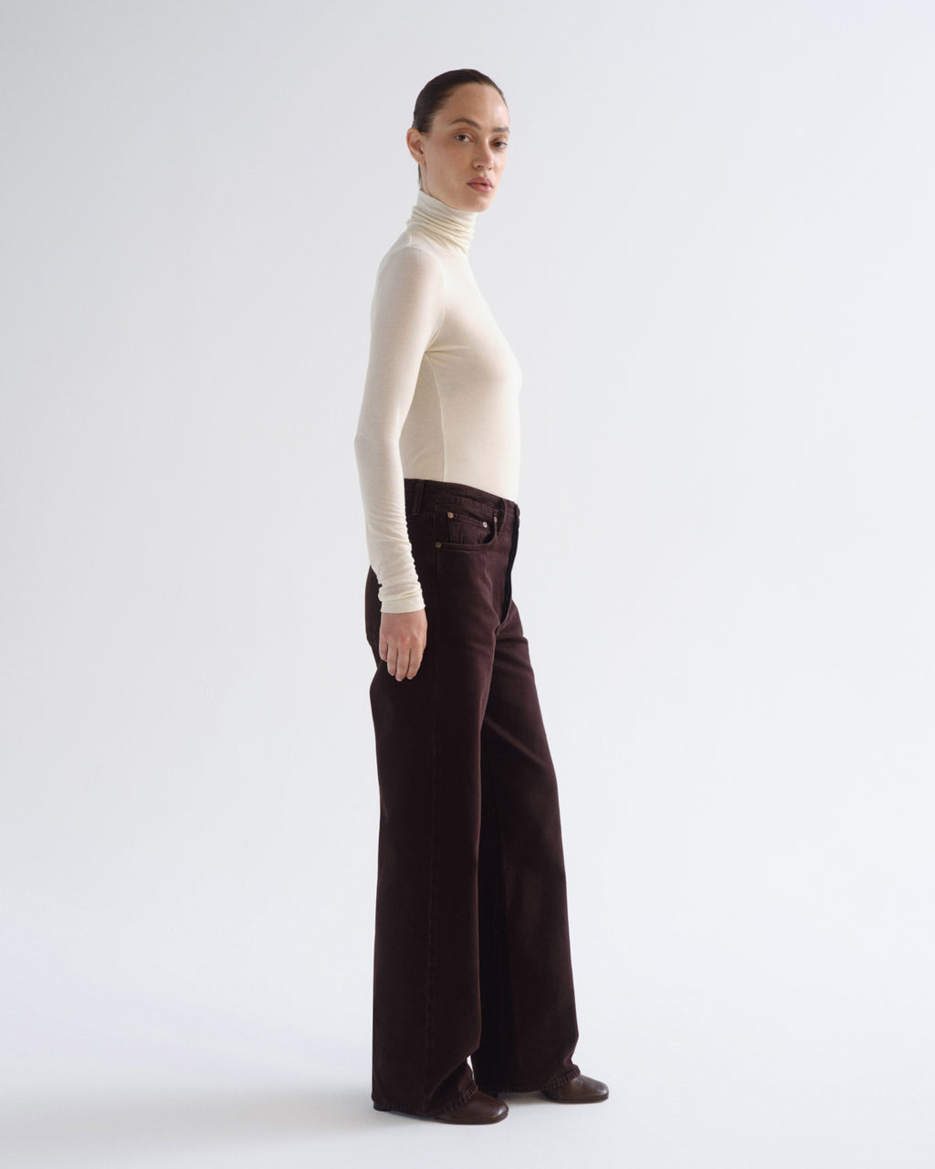 A model standing in profile wearing a cream turtleneck top and dark brown wide-leg trousers against a light gray studio background.