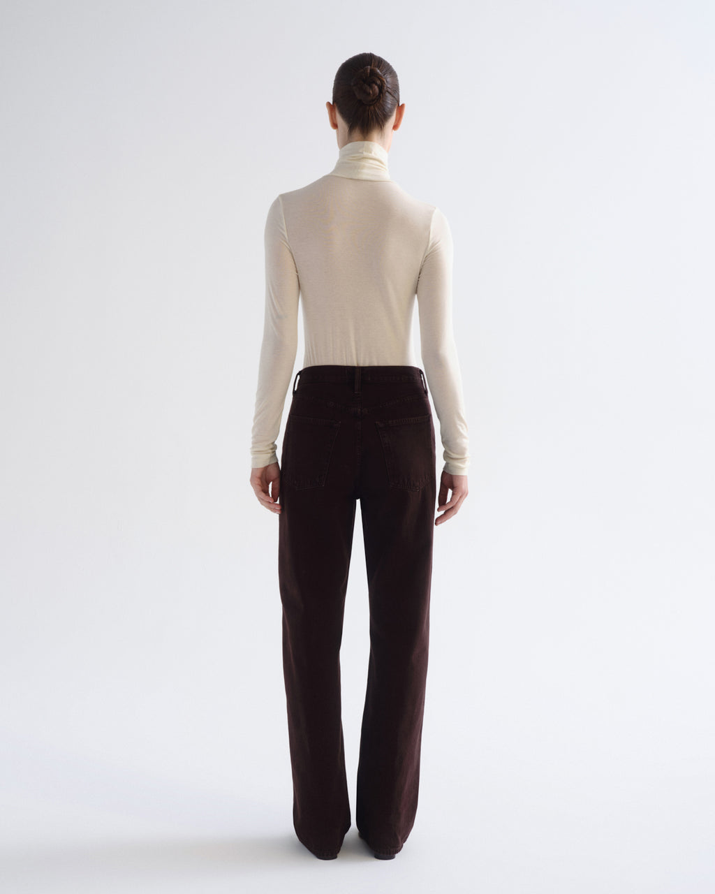 A model shown from the back wearing a cream turtleneck top and dark brown wide-leg trousers, photographed against a seamless light gray studio background.