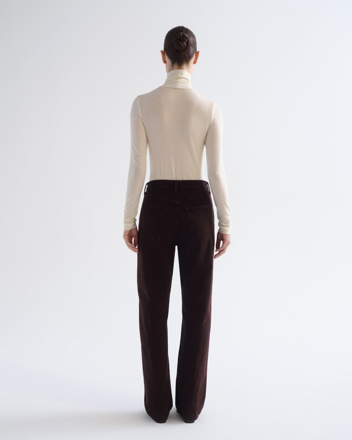 A model shown from the back wearing a cream turtleneck top and dark brown wide-leg trousers, photographed against a seamless light gray studio background.