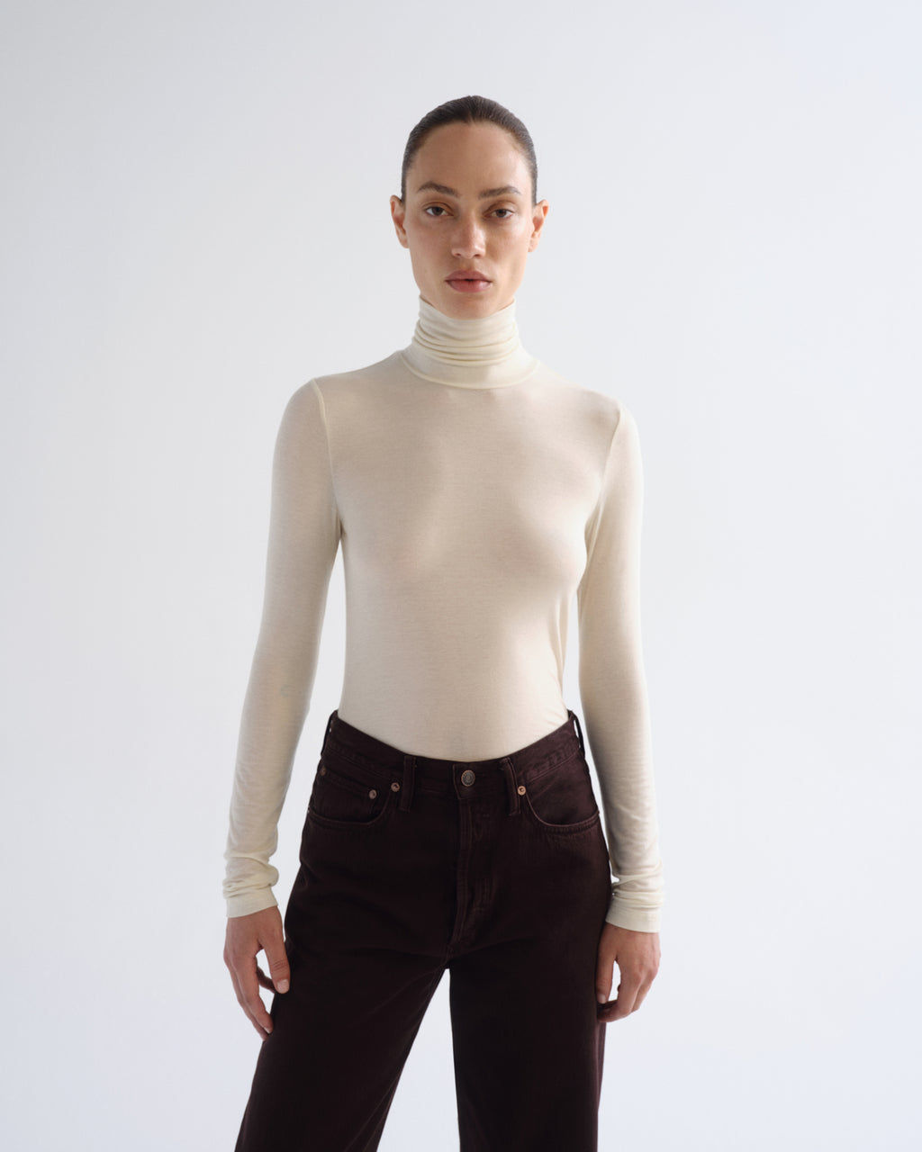 A model wearing a fitted, long-sleeve cream turtleneck top and dark brown corduroy pants, standing against a light gray studio background.