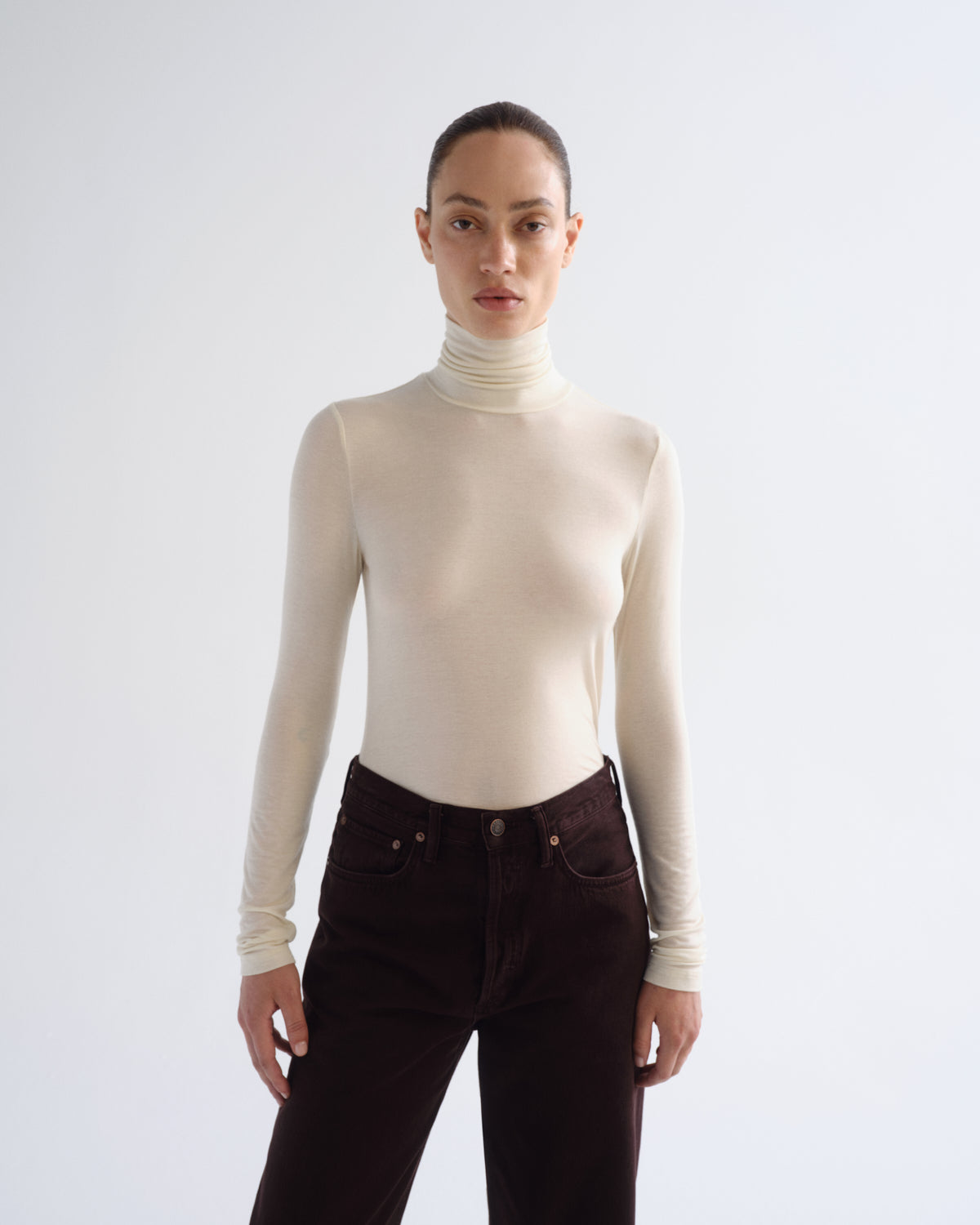 A model wearing a fitted, long-sleeve cream turtleneck top and dark brown corduroy pants, standing against a light gray studio background.