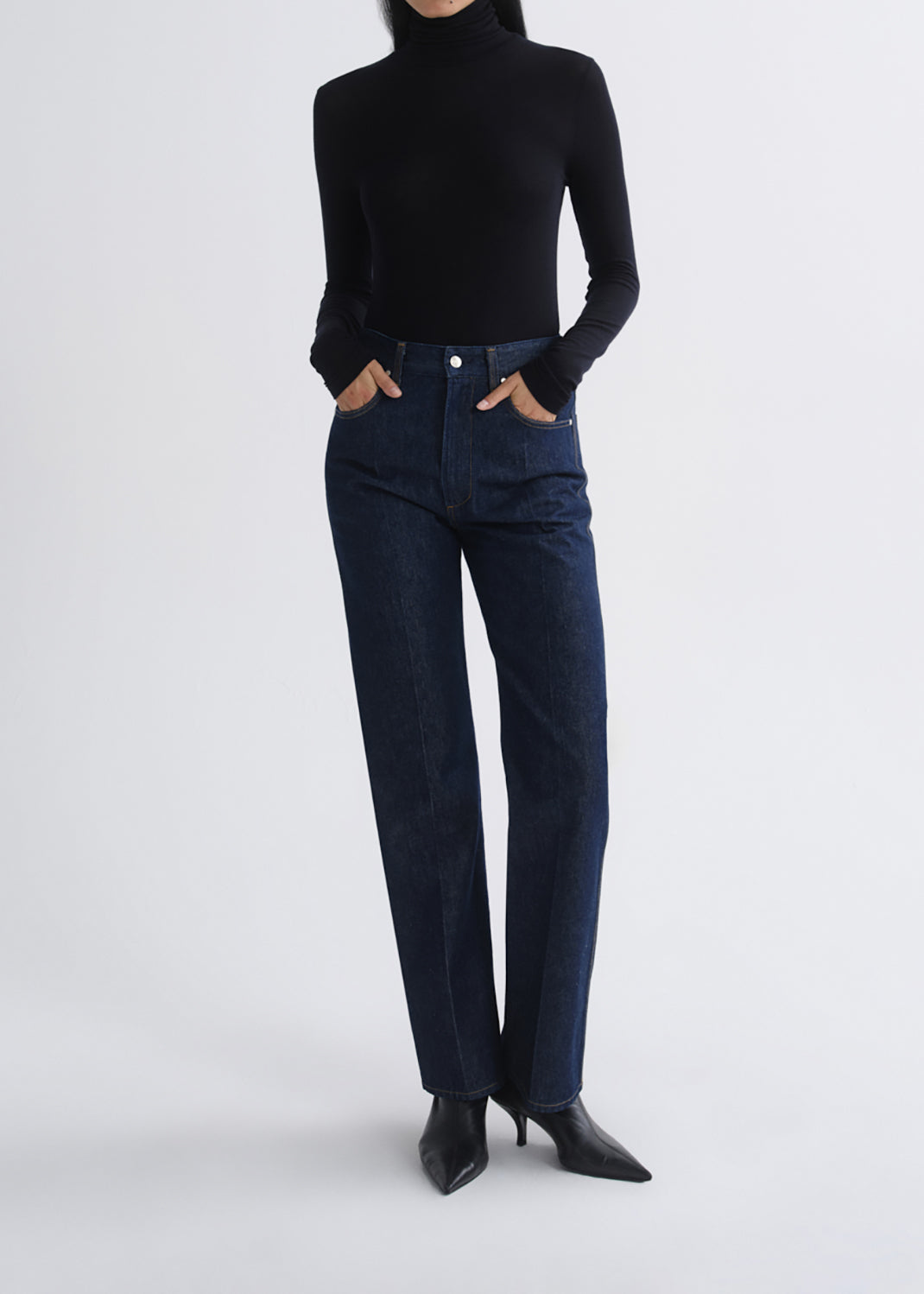 A model wearing dark blue straight-leg denim jeans, a black long-sleeve turtleneck top, and black pointed-toe heeled boots, standing against a grey background.