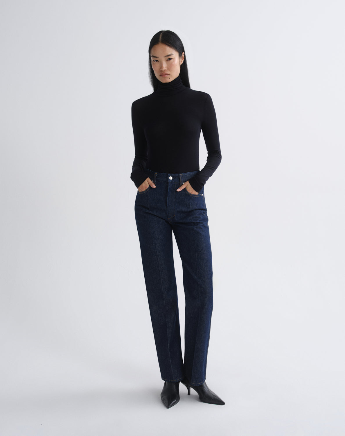 A model stands against a light gray studio background wearing a fitted black long-sleeve turtleneck sweater, dark blue straight-leg jeans, and black pointed-toe heels.