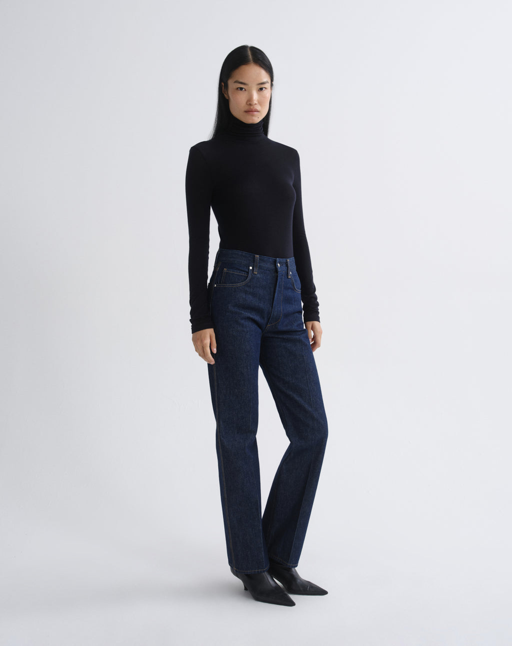 A model stands against a light gray studio background wearing a black long-sleeve turtleneck sweater, dark blue straight-leg jeans, and black pointed-toe flats.