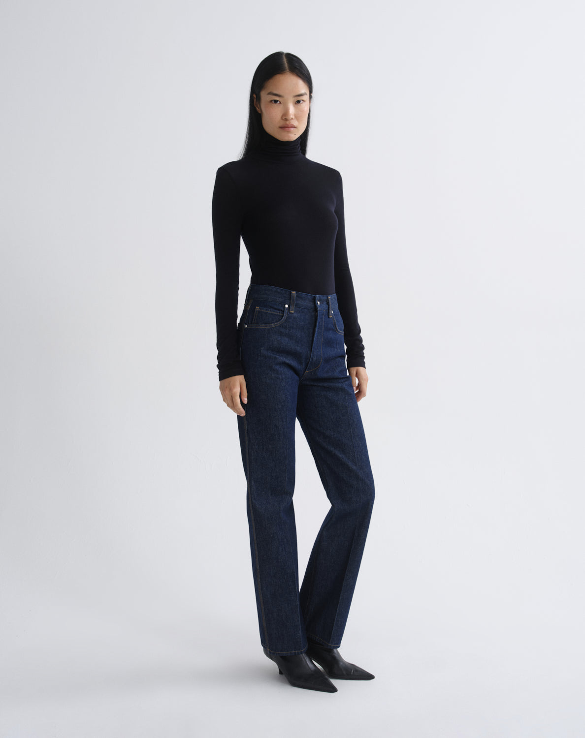 A model stands against a light gray studio background wearing a black long-sleeve turtleneck sweater, dark blue straight-leg jeans, and black pointed-toe flats.