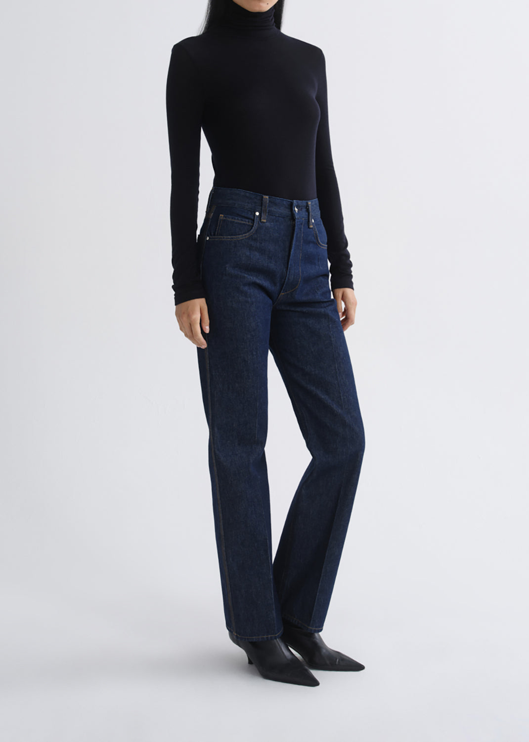 A model wearing dark blue high-rise straight-leg jeans, a black long-sleeve turtleneck, and black pointed-toe ankle boots, standing against a grey background.