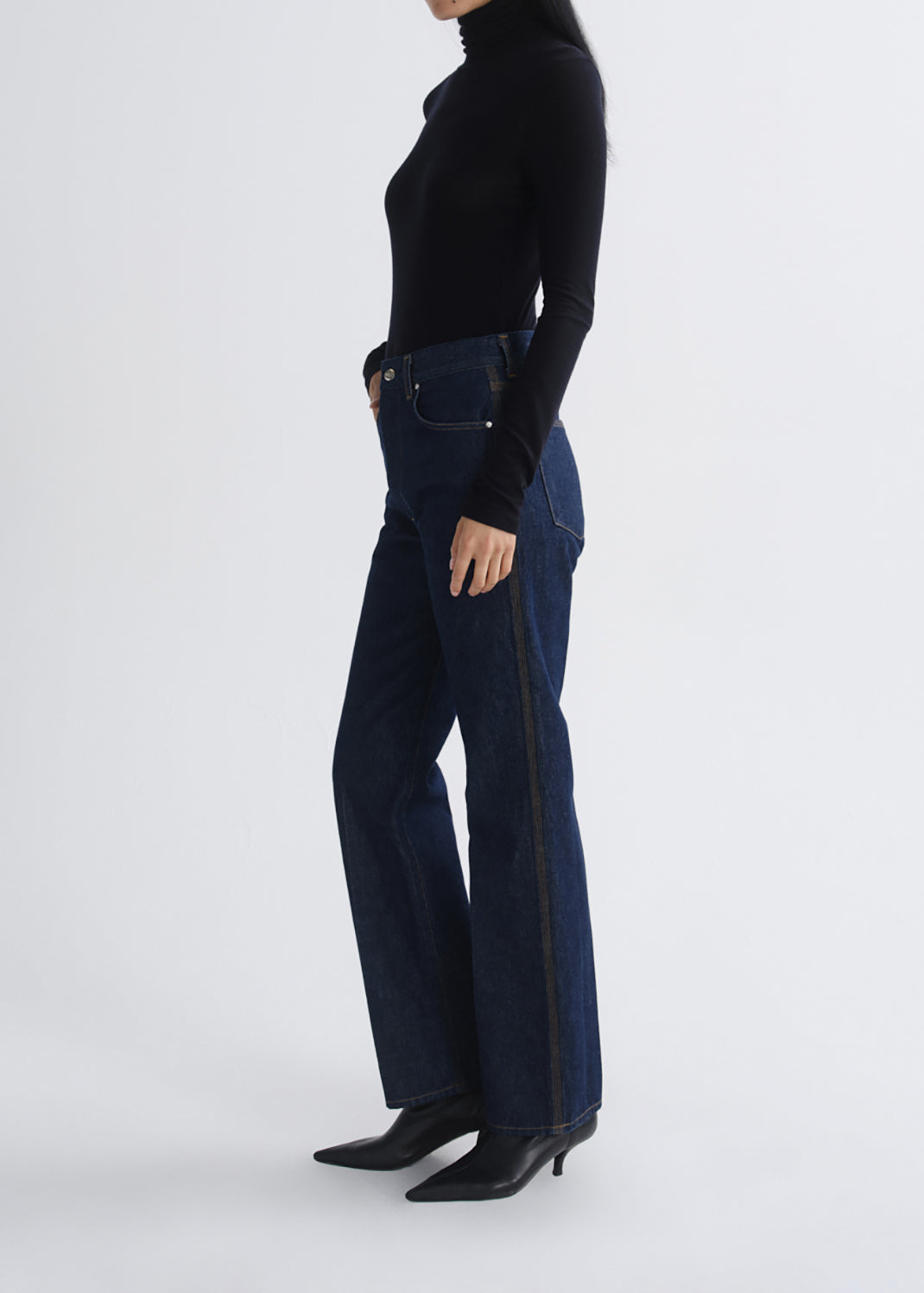 A model shown from the side wearing dark blue flared jeans with a black turtleneck and black pointed-toe heeled boots, against a grey background.