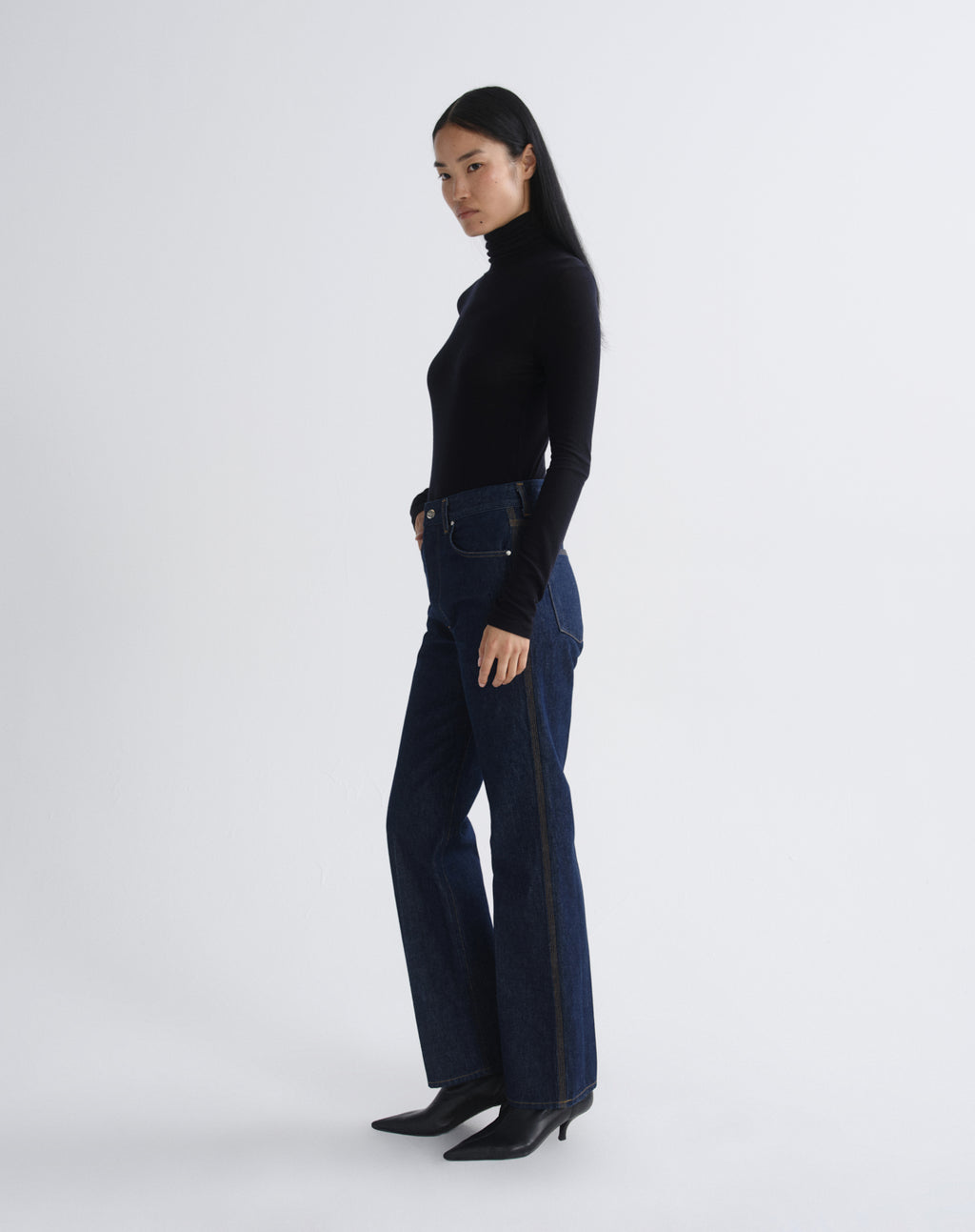 A model is shown in profile wearing a black long-sleeve turtleneck top, dark blue high-waisted flare jeans with a black side stripe, and black pointed-toe heels, against a light gray studio background.
