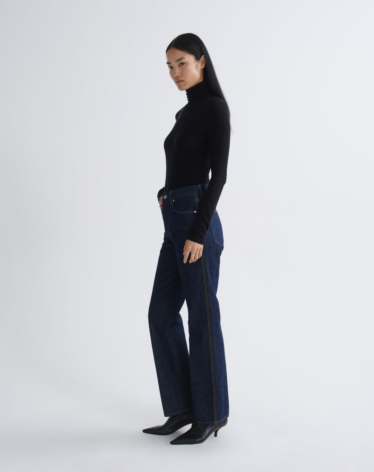 A model is shown in profile wearing a black long-sleeve turtleneck top, dark blue high-waisted flare jeans with a black side stripe, and black pointed-toe heels, against a light gray studio background.
