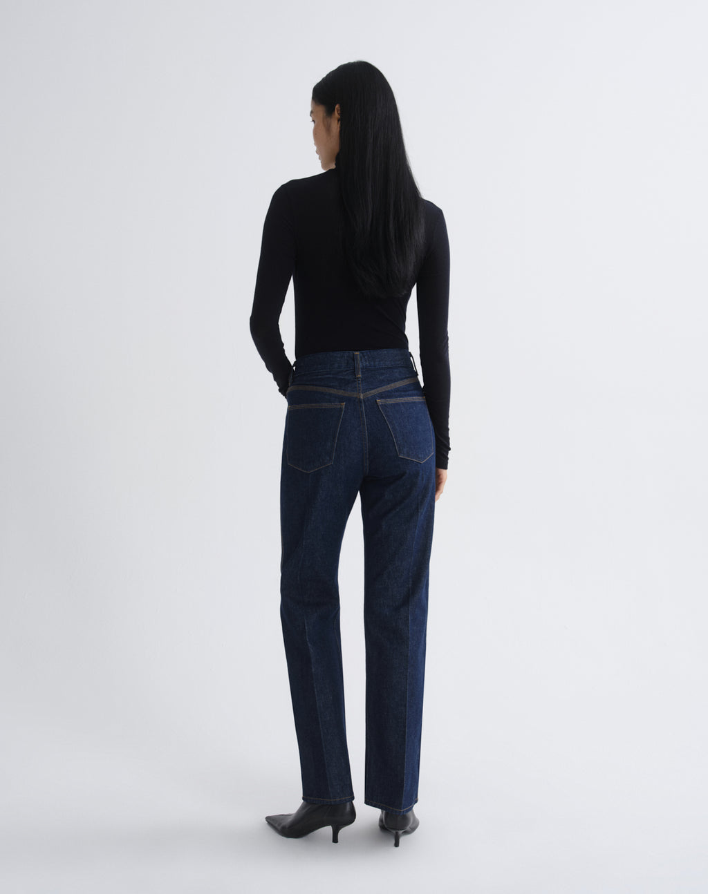 A woman is shown from behind wearing dark blue straight-leg jeans, a black long-sleeve top, and black pointed-toe heels against a light gray studio background.