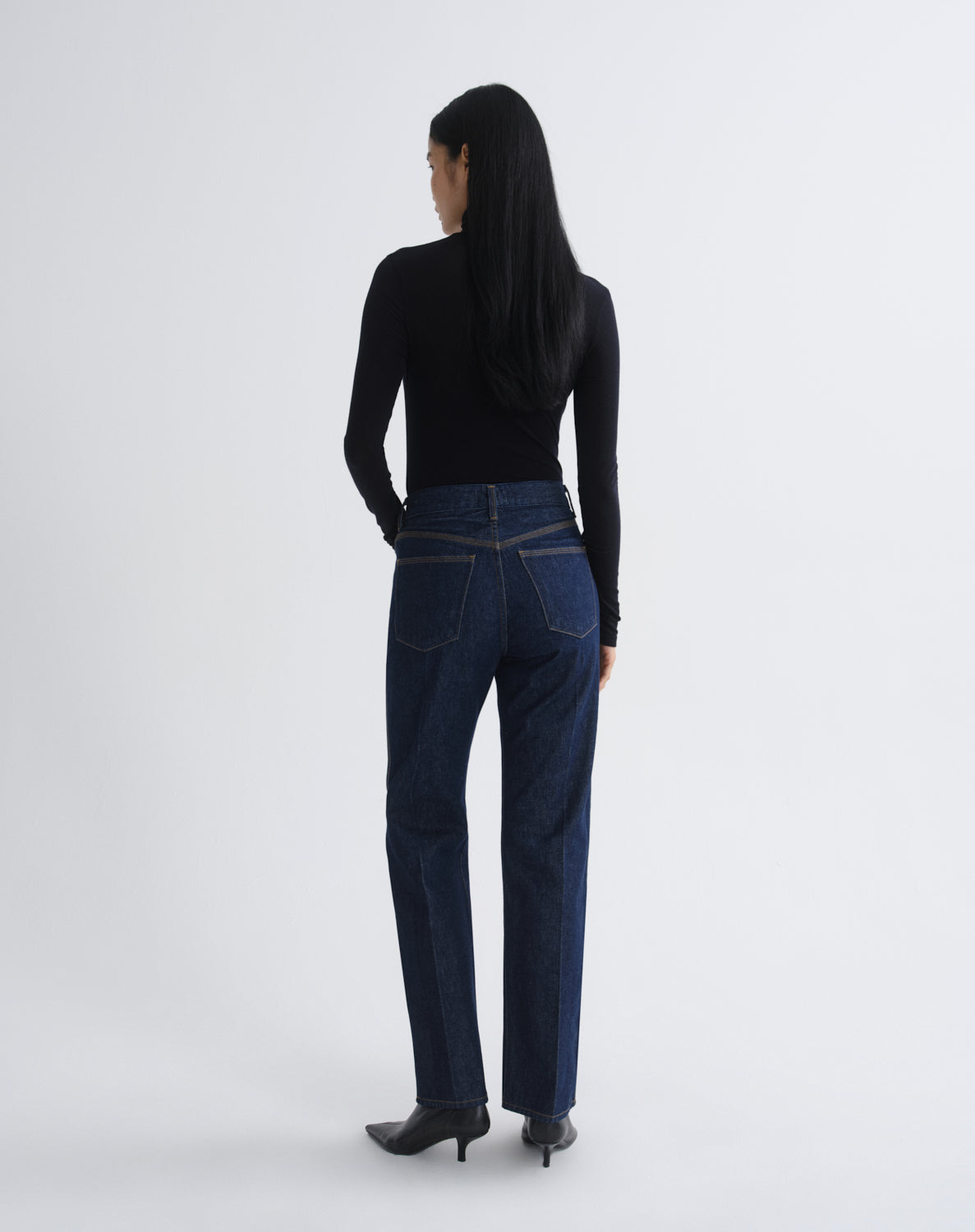 A woman is shown from behind wearing dark blue straight-leg jeans, a black long-sleeve top, and black pointed-toe heels against a light gray studio background.