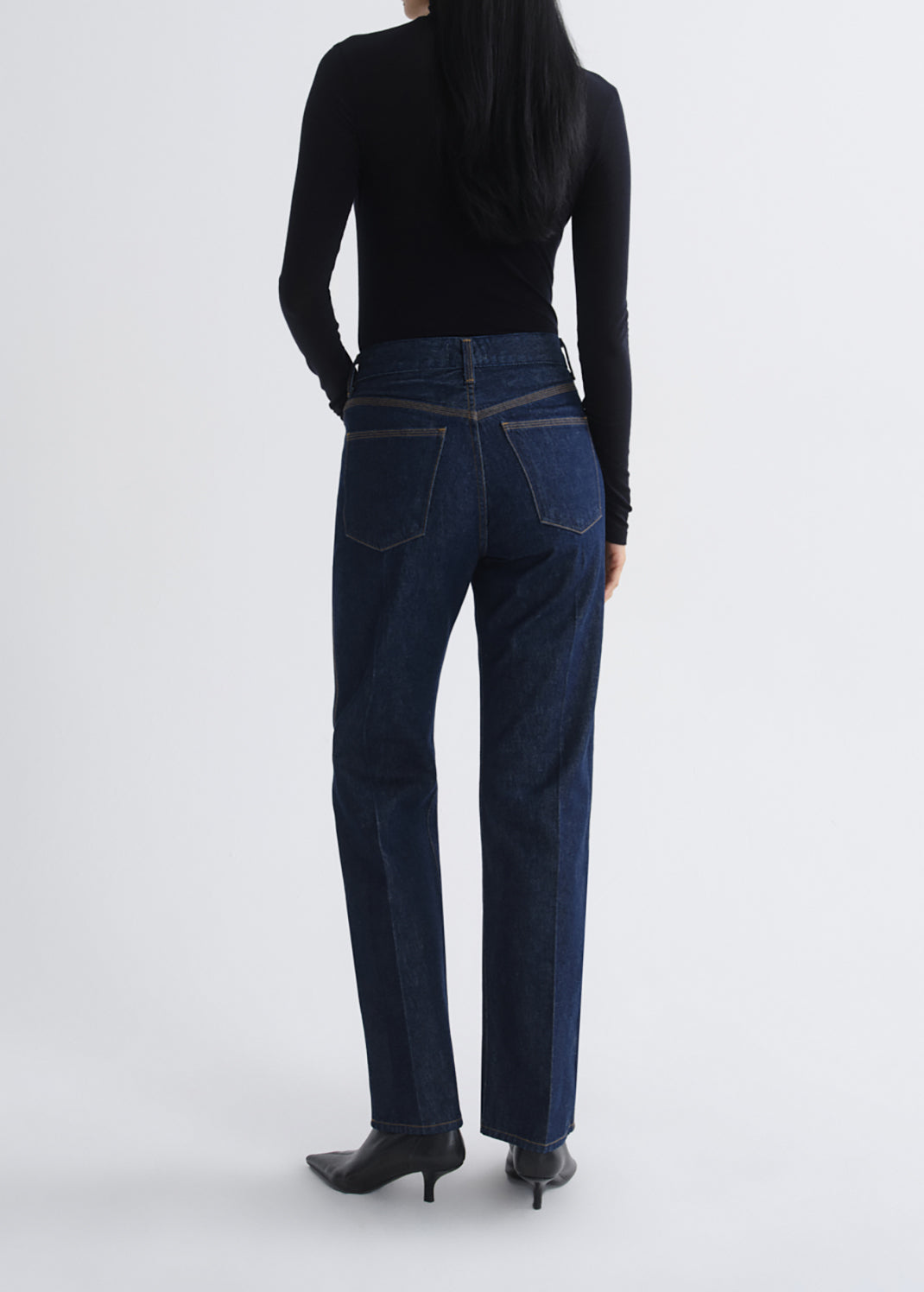 A model viewed from the back wearing dark blue straight-leg jeans, a black long-sleeve top, and black pointed-toe heels against a grey background.