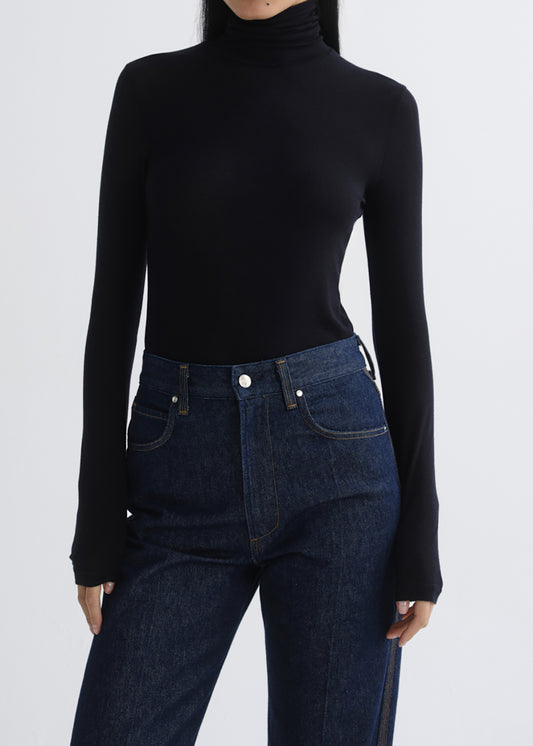 A model wearing a black long-sleeve turtleneck top and dark blue jeans, cropped from the neck down against a grey background.