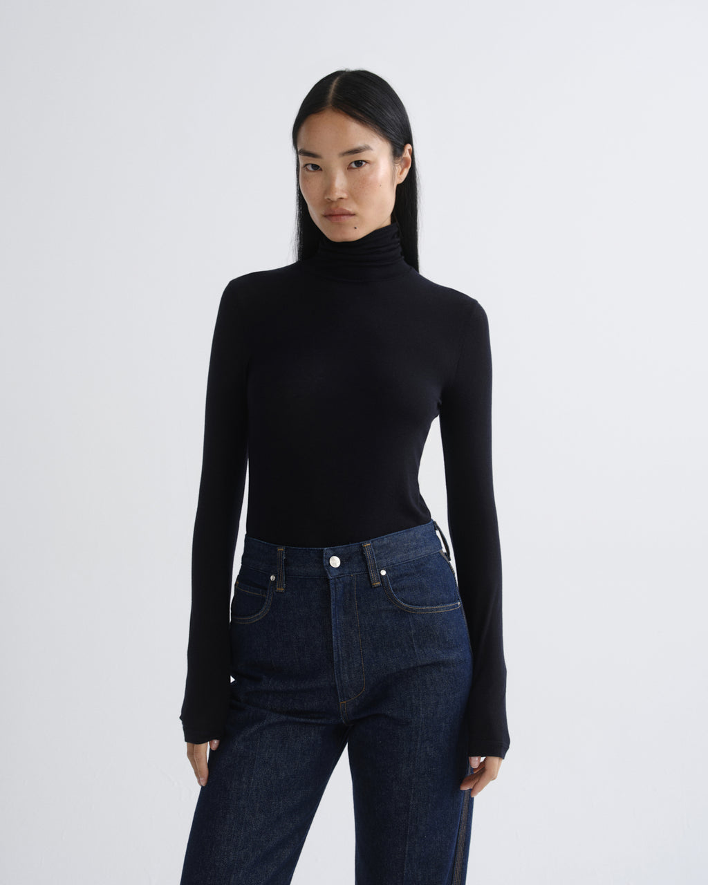 A model wearing a black long-sleeve turtleneck top and dark blue jeans, standing against a light gray studio background.
