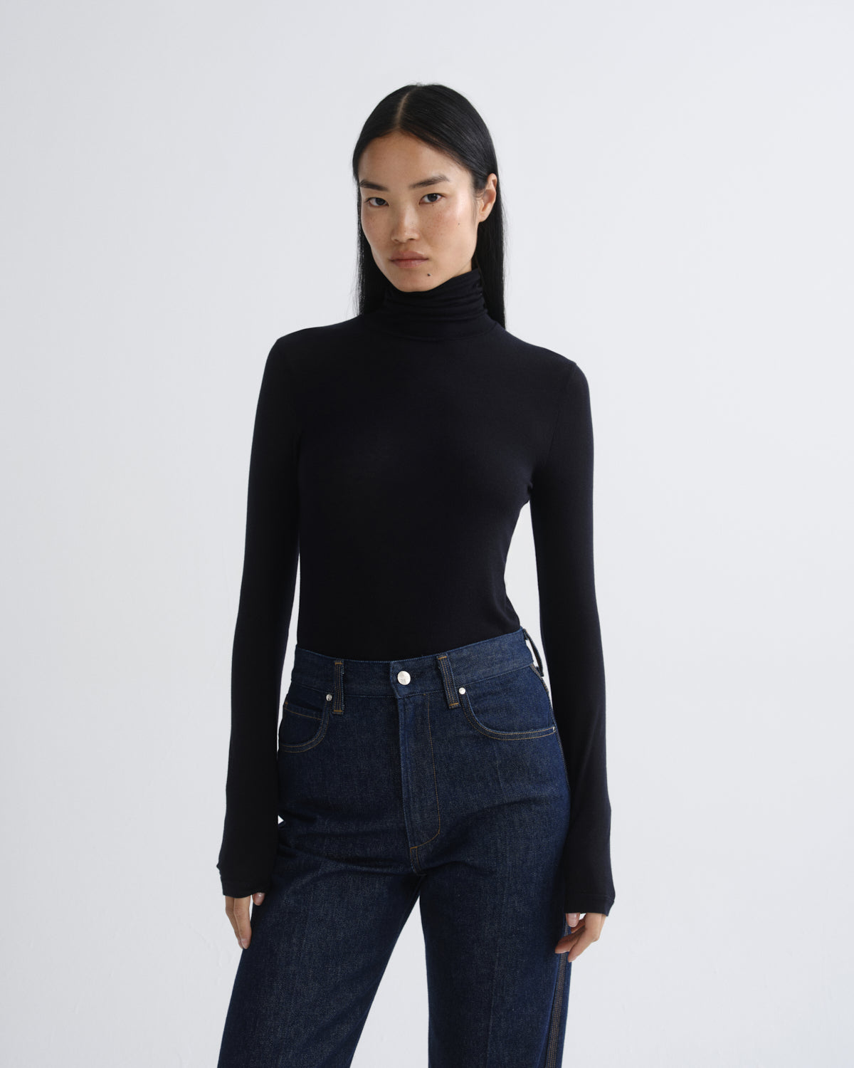A model wearing a black long-sleeve turtleneck top and dark blue jeans, standing against a light gray studio background.