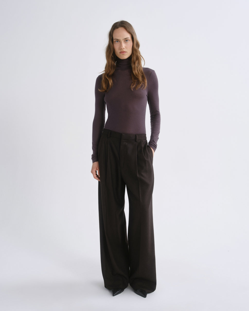 A woman modeling a dark plum long-sleeve mock neck top paired with dark brown wide-leg trousers, standing against a light gray studio background.