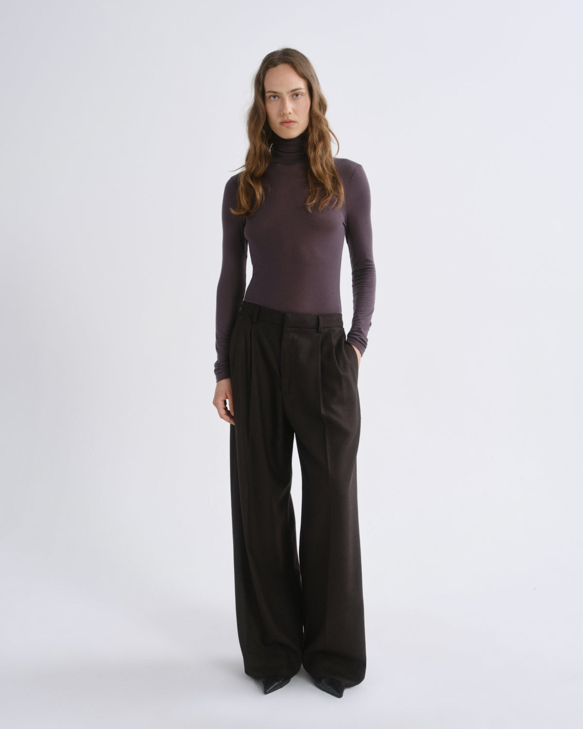 A woman modeling a dark plum long-sleeve mock neck top paired with dark brown wide-leg trousers, standing against a light gray studio background.
