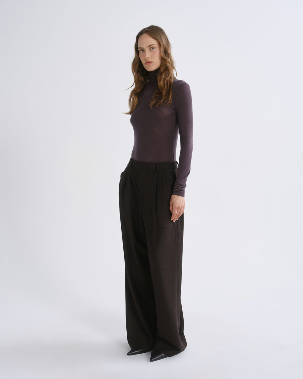 A woman modeling a dark plum long-sleeve mock neck top paired with wide-leg black trousers, standing against a light gray studio background.