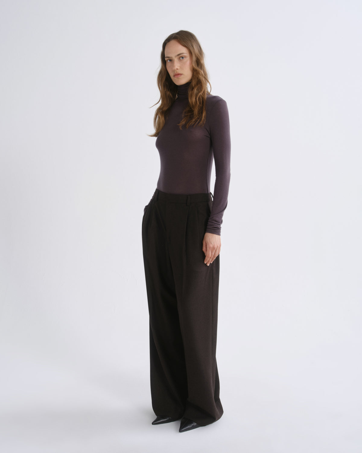 A woman modeling a dark plum long-sleeve mock neck top paired with wide-leg black trousers, standing against a light gray studio background.