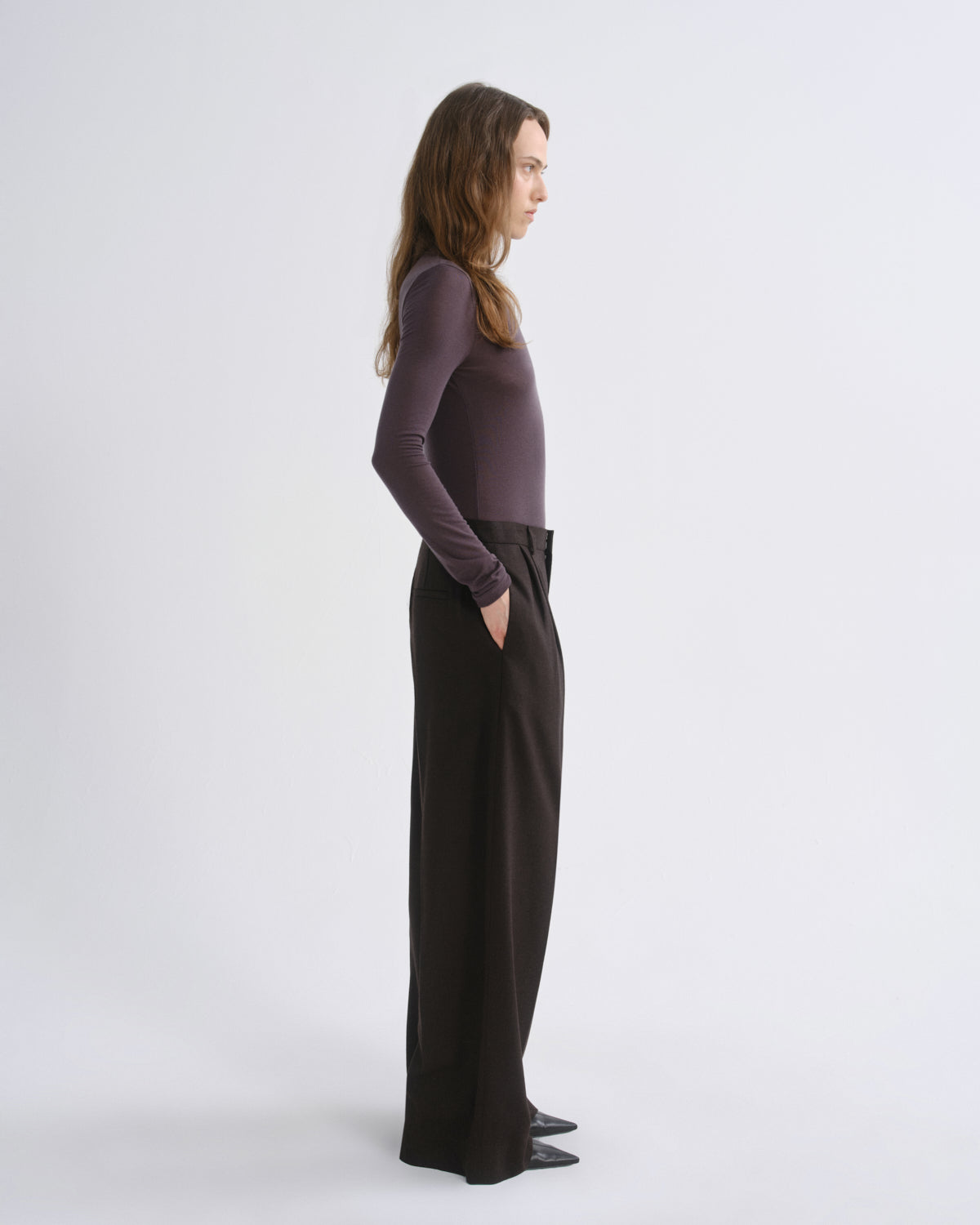 A woman stands in profile wearing a fitted long-sleeve top in a muted plum-purple and wide-leg dark trousers, photographed against a clean light gray studio background.