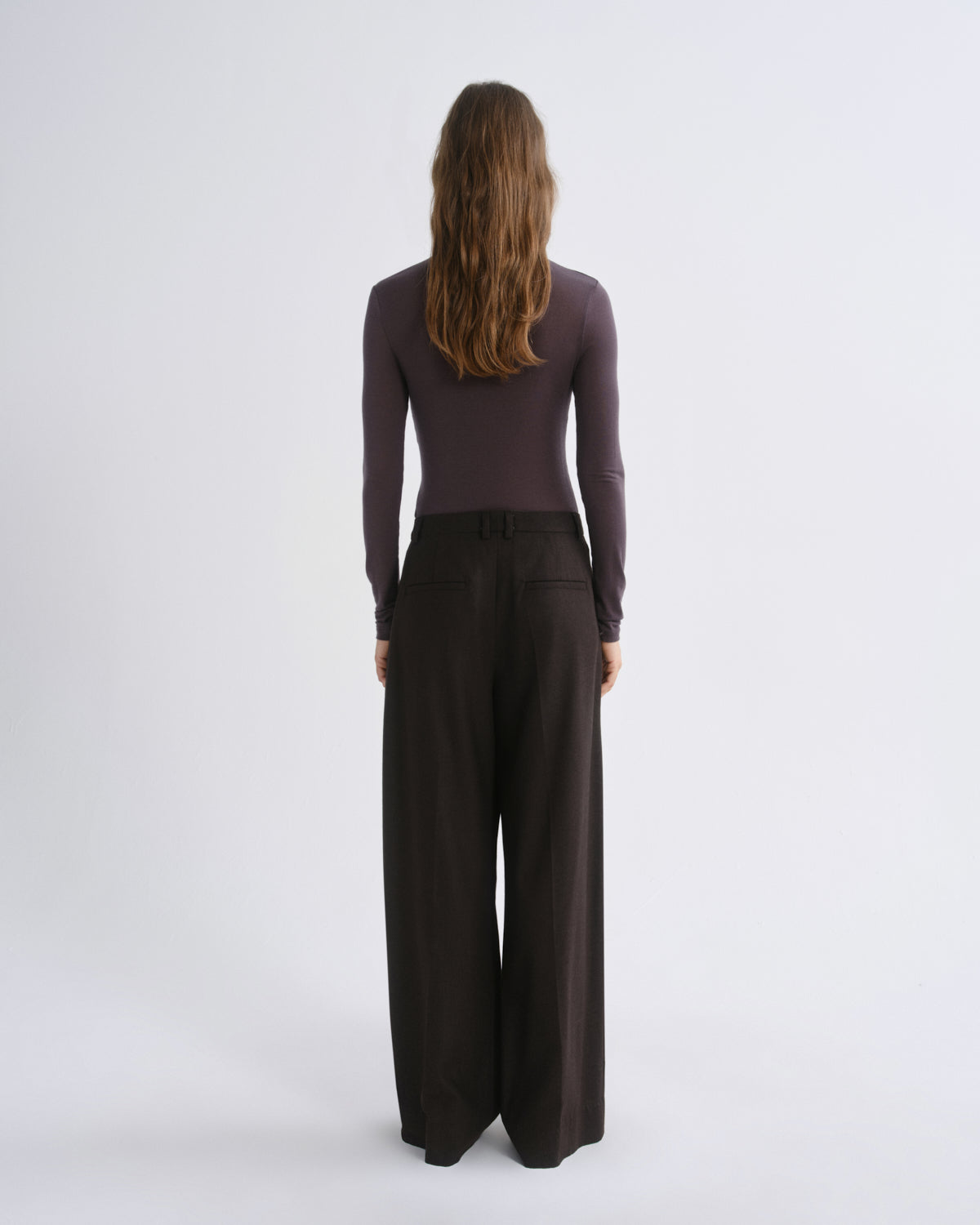 A woman viewed from behind wearing a dark plum long-sleeve top and dark brown wide-leg trousers against a light gray studio background.