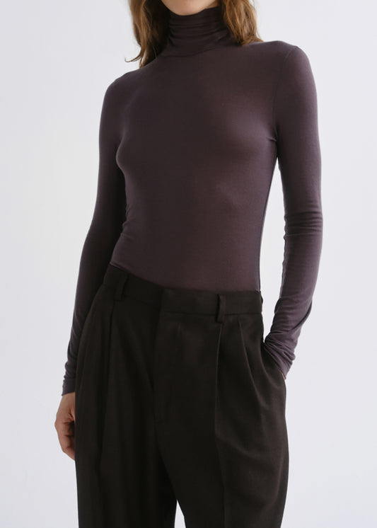 A model wearing a dark plum long-sleeve mock neck top paired with dark brown pleated trousers, shown from the neck down against a grey background.