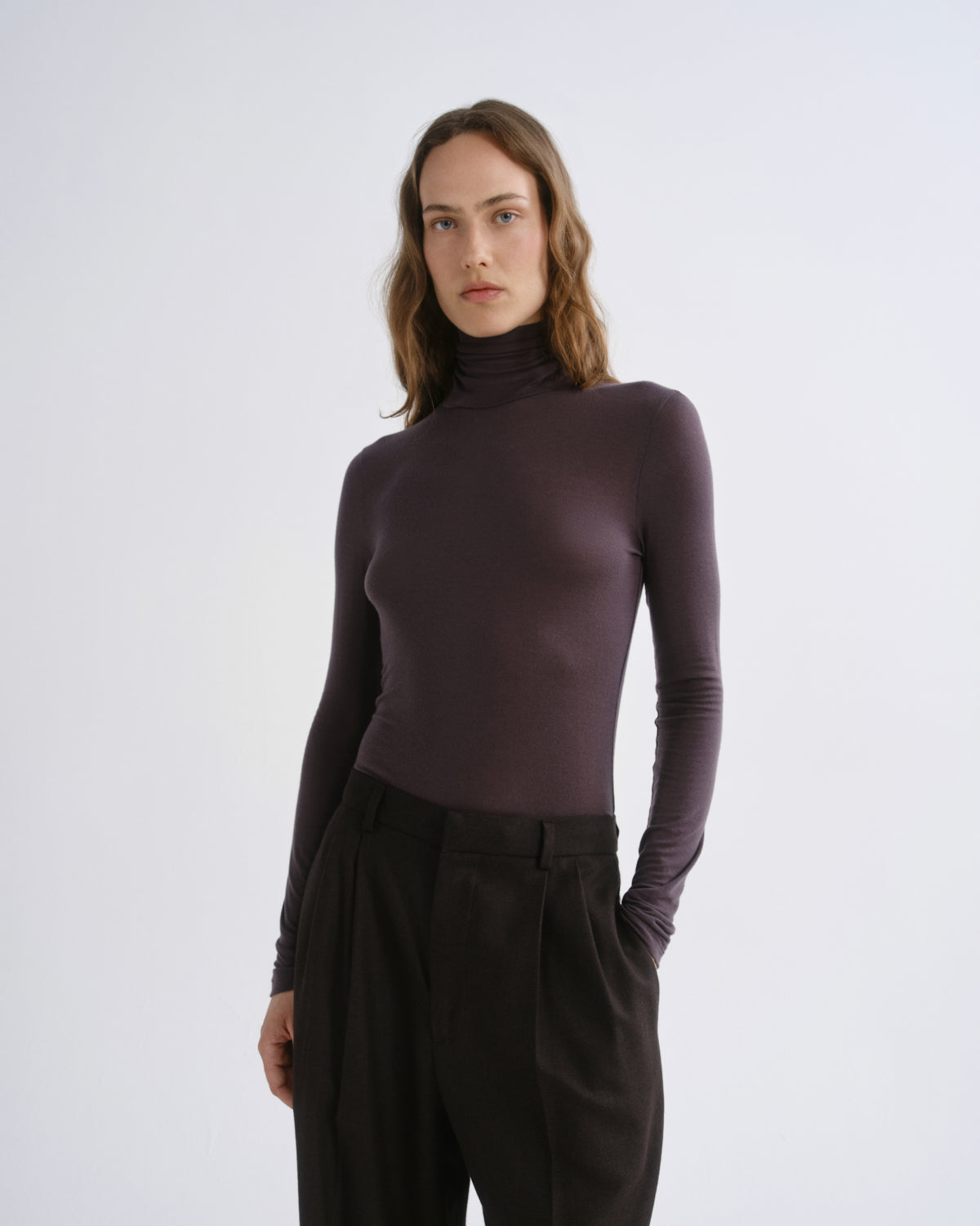 A woman modeling a fitted, long-sleeve mock neck turtleneck top in a deep plum purple color, paired with black trousers, against a plain light gray studio background.