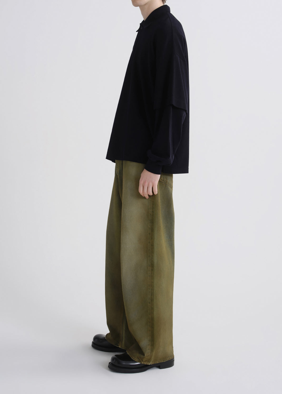 A model shown from the side wearing a black long-sleeve shirt and wide-leg olive green trousers, paired with black leather shoes, against a grey background.