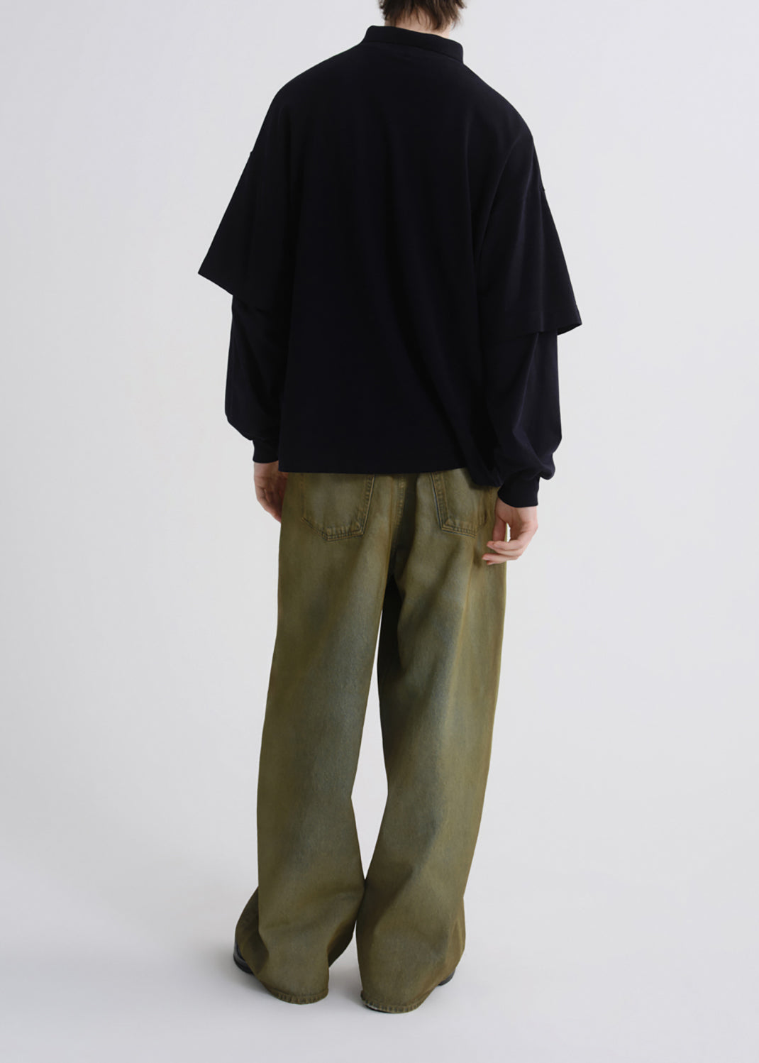 A model shown from the back wearing a black long-sleeve top with layered sleeves and olive green wide-leg trousers, photographed against a grey background.