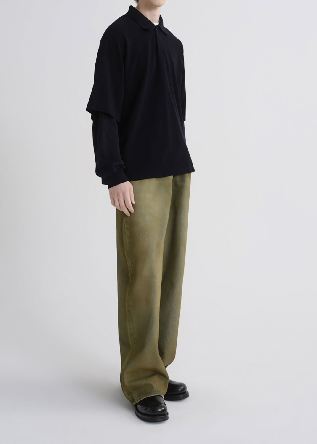 A model wearing a black long-sleeve collared shirt with layered sleeves and wide-leg olive green trousers, paired with black leather shoes, against a grey background.
