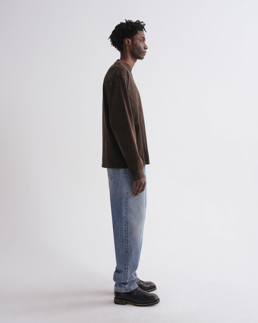 A man standing in profile wearing a dark brown long-sleeve crewneck shirt, light blue jeans, and black shoes against a plain light gray studio background.