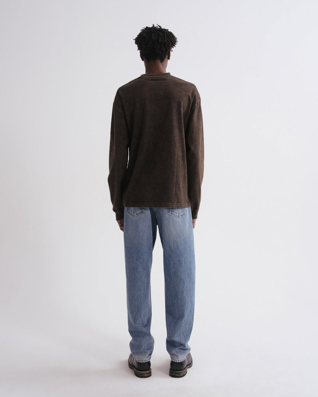 A man viewed from behind wearing a dark brown long-sleeve knit sweater and light blue jeans, standing against a plain light gray studio background.