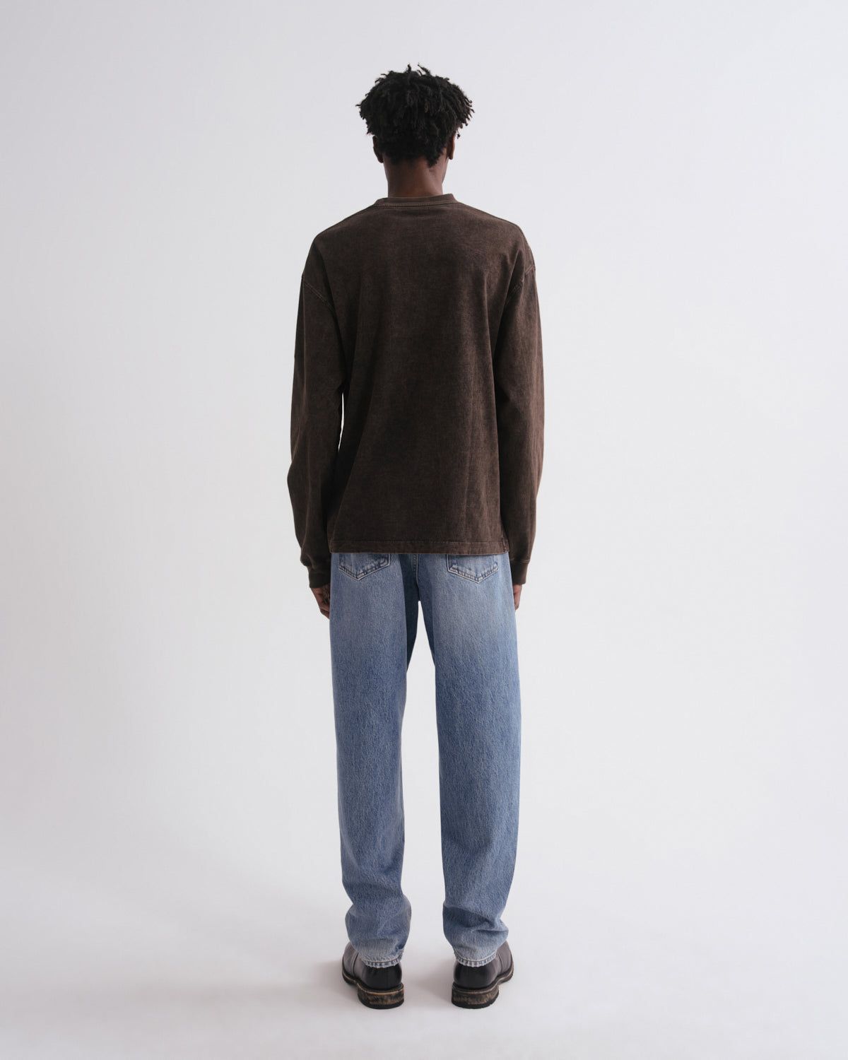 A man viewed from behind wearing a dark brown long-sleeve knit sweater and light blue jeans, standing against a plain light gray studio background.