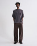 A man stands against a grey background wearing an oversized dark charcoal grey t-shirt and loose dark brown pants.