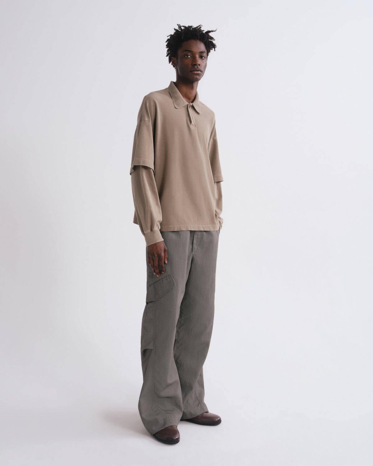 A model stands against a light gray studio background wearing a beige long-sleeve collared shirt with gathered sleeves and wide-leg taupe-gray trousers.