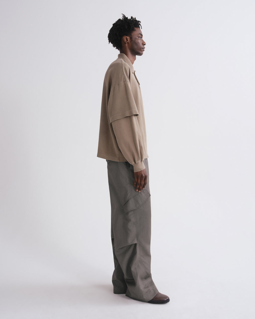 A man stands in profile wearing a beige oversized long-sleeve shirt and wide-leg gray trousers against a plain light gray studio background.