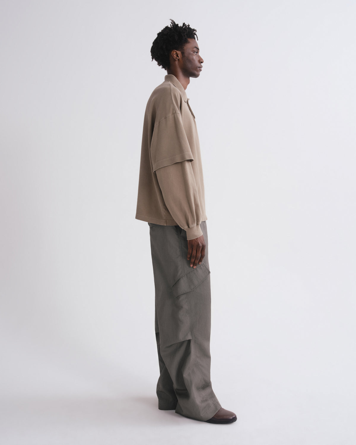 A man stands in profile wearing a beige oversized long-sleeve shirt and wide-leg gray trousers against a plain light gray studio background.