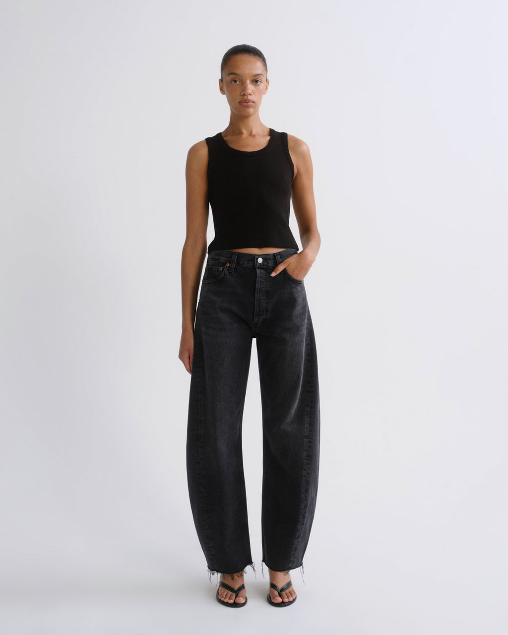 A model stands against a light gray studio background wearing a black ribbed sleeveless crop top and wide-leg dark gray jeans with fringe hems, paired with black sandals.