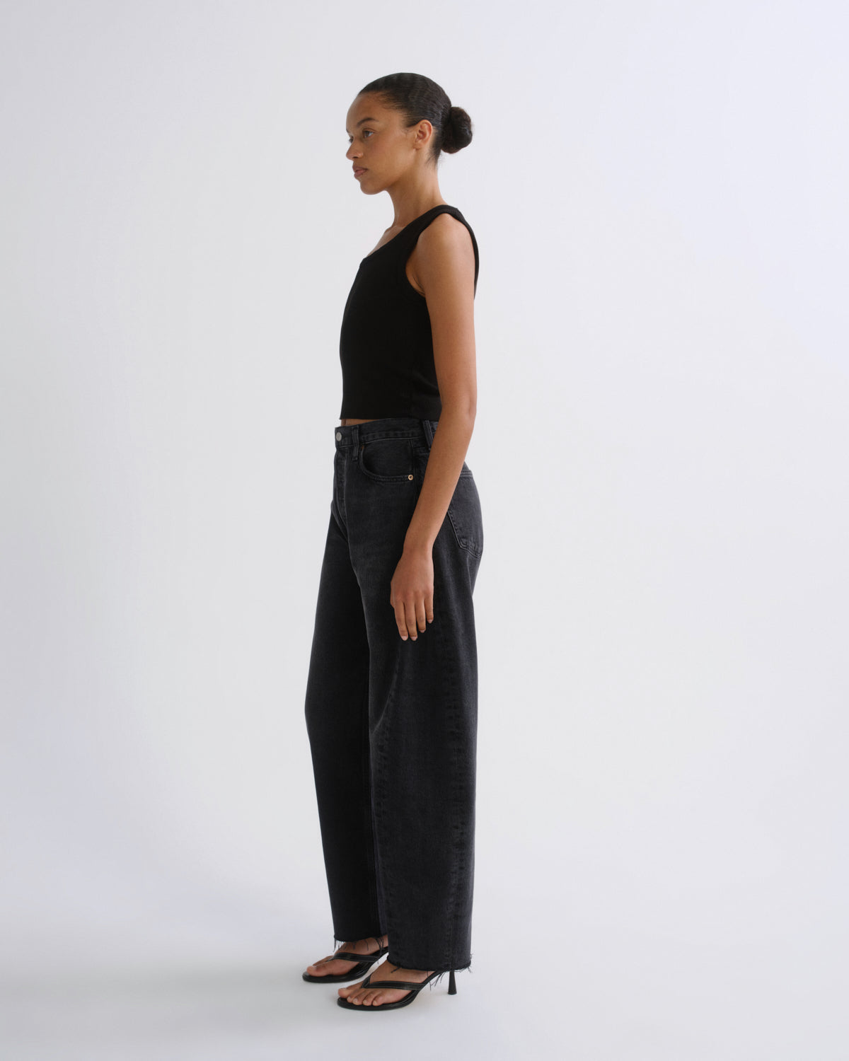 A woman stands in profile wearing a black sleeveless top, dark wide-leg jeans, and black heeled sandals against a plain light gray studio background.