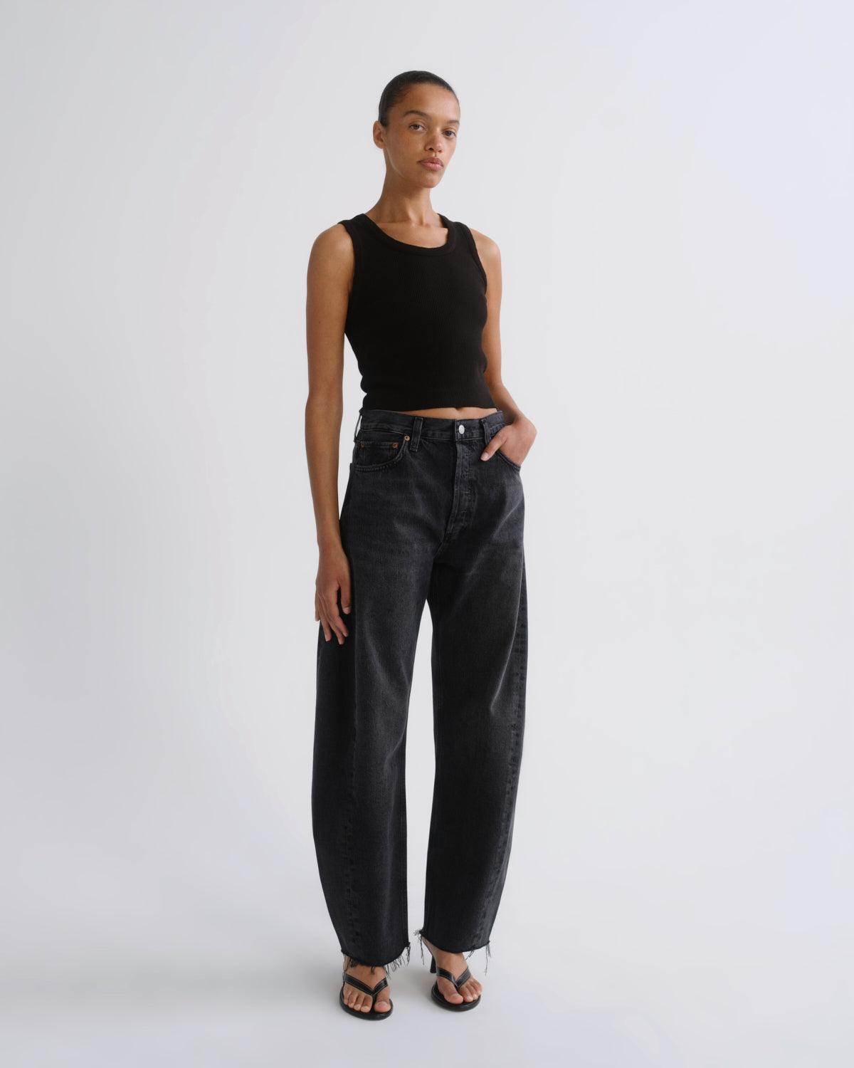 A model stands against a light gray studio background wearing a black sleeveless crop top, dark charcoal wide-leg jeans, and black sandals.