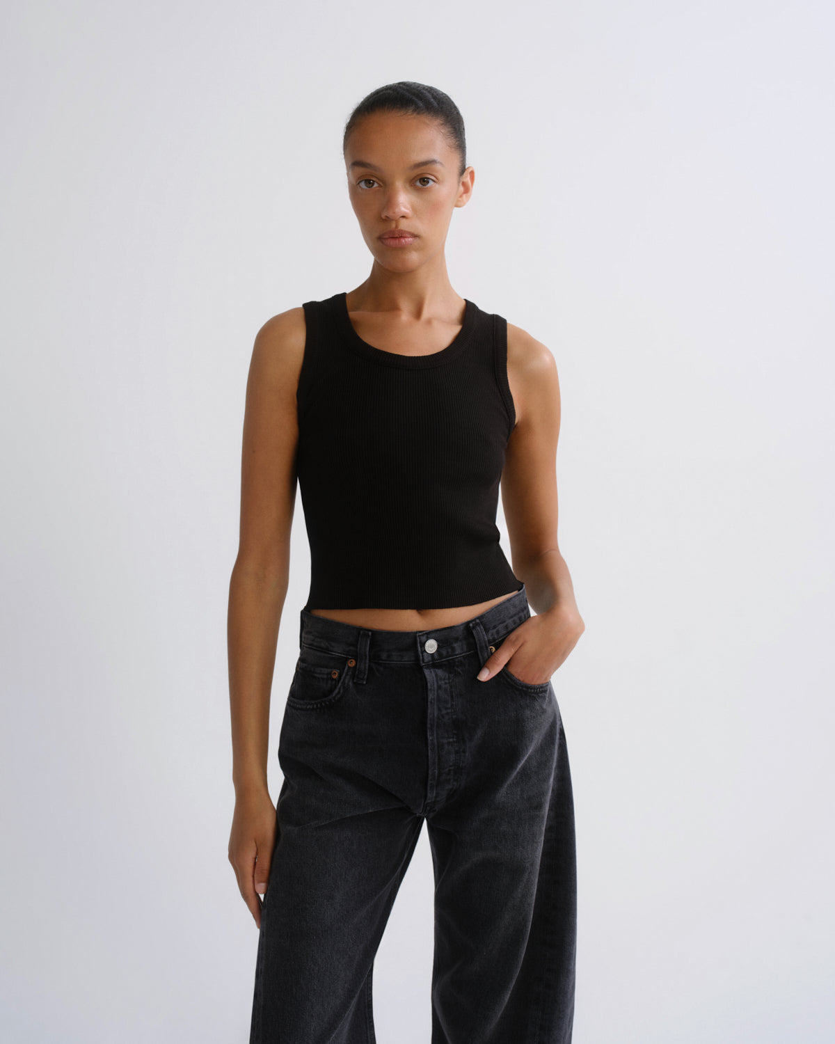 A model wearing a black ribbed sleeveless crop top and dark gray wide-leg jeans, standing against a light gray studio background.