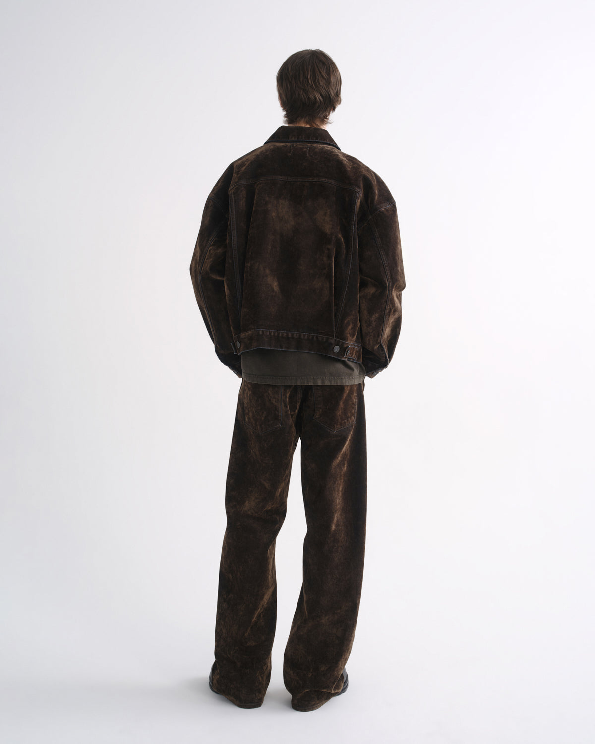Person wearing a dark brown denim jacket with matching dark brown pants and black shoes, back view, on a grey background